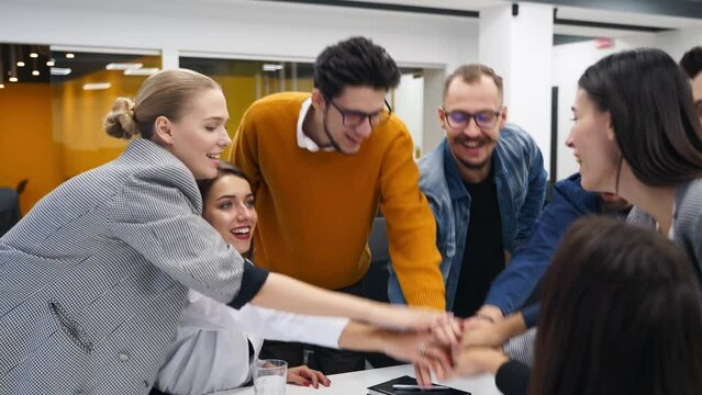 Business people stack hands in pile over meeting table, cheer together in corporate teambuilding event in office. Coworkers help, support teamwork. Colleagues join to celebrate success. Unity concept.