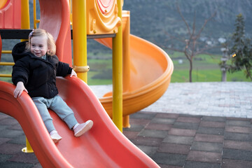 Happy blonde caucasian pretty girl, toddler wearing warm clothes sliding on playground,having...
