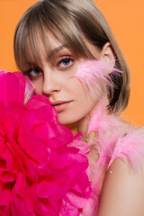 young woman with decorative beads in makeup and feathers on cheeks near pink flower isolated on orange