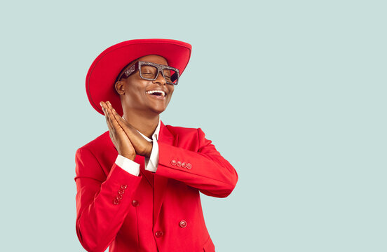Funny African man in trendy outfit dancing in studio. Happy young Tanzanian guy wearing funky red suit, hat and disco party glasses dancing isolated on blue copy space background. Fashion concept