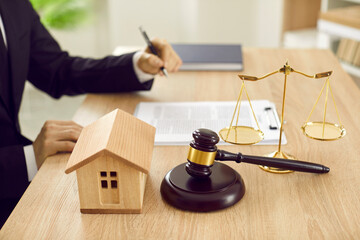 Attorney sitting at desk with scales of justice, gavel and small wooden toy house, working with documents, signing contract agreements. Real estate law, foreclosed property, lawyer services concept - obrazy, fototapety, plakaty
