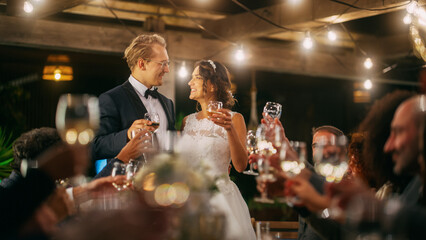 Beautiful Bride and Groom Celebrate Wedding at an Evening Reception Party. Newlyweds Propose a Toast to Happy Marriage, Standing at a Dinner Table with Best Multiethnic Diverse Friends.