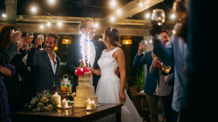 Beautiful Bride and Groom Celebrate Wedding at an Evening Reception Party with Multiethnic Friends. Newlyweds Propose a Toast to Happy Marriage, Standing at a Dinner Table with Cake with Sparklers.