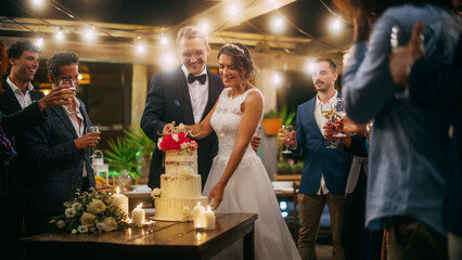 Beautiful Bride and Groom Celebrate Wedding at an Evening Reception Party with Multiethnic Friends. Married Couple Standing at a Dinner Table, Cutting Wedding Cake.