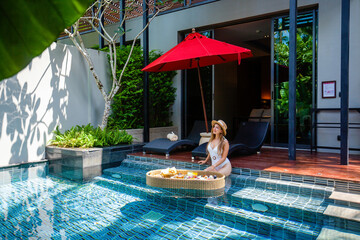 Romantic girl in swimwear relaxing and eating floating breakfast on rattan tray in pool on luxury...