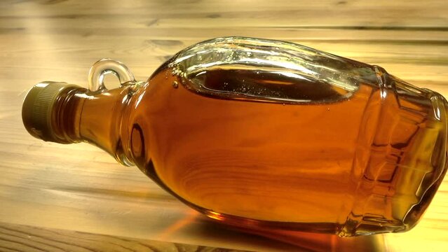 Closeup of a bottle of  translucent amber Canadian maple syrup, rocking on its side, on a pine worktop / counter.