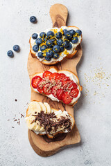 Fruity vegetarian toasts for breakfast on gray background. Bread slices with ricotta, berries, banana, chocolate and seeds.