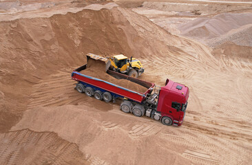 Wheel loader load sand in tipper truck in open pit. Front end loader on earthworks in open-pit mining. Heavy machinery on developing opencast. Dump truck sand transports from quarry, aerial view.