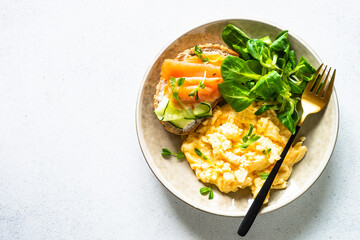 Scrambled eggs, sandwich with cream cheese and salmon and salad leaves at white stone table.