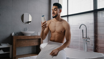 Arab Indian guy muscular naked sexy unshaven man wears white bath towel on hips sits in bathroom getting ready for bathing washing sniffing shower gel morning refreshment with natural male cosmetics