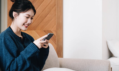 Smiling young asian woman using mobile phone while sitting on a couch at home with laptop computer