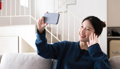 Smiling young asian woman using mobile phone while sitting on a couch at home with laptop computer - obrazy, fototapety, plakaty
