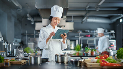 World Famous Restaurant: Asian Female Chef Cooking Delicious and Authentic Food, Uses Digital Tablet Computer While Working in a Modern Professional Kitchen. Preparing gourmet organic Dishes - obrazy, fototapety, plakaty