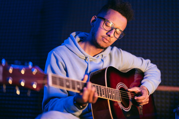 Young afro musician wearing glasses playing acoustic guitar, on dark background