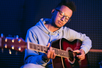 Obraz premium Young afro musician wearing glasses playing acoustic guitar, on dark background