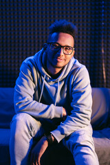 Dark skinned young man with a beaming smile, wears a gray parka, and glasses on dark background splashed by blue light