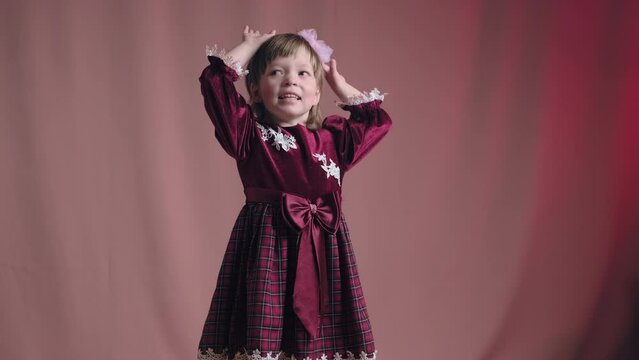 Cute little girl in an vintage burgundy dress is dancing, rejoicing and making funny faces. The concept of children theater productions and developing acting circles. Pink background