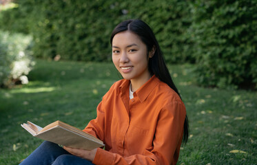 Beautiful smiling Vietnamese student studying, reading book looking at camera sitting in park....