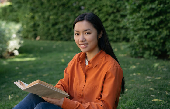 Beautiful smiling Vietnamese student studying, reading book looking at camera sitting in park. Education concept