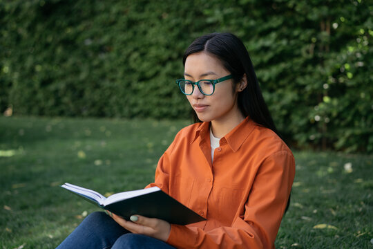 Portrait of pensive asian woman wearing casual clothing and stylish eyeglasses reading book sitting in park. Education concept   
