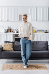 full length of stylish woman posing with hands in pockets of jeans near couch in open plan kitchen