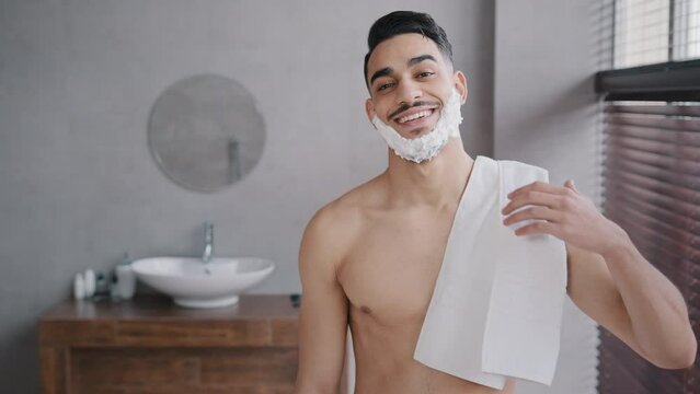 Funny male portrait in bathroom hispanic indian arabian naked man bearded guy posing smiling with white soapy foam on beard shaving gel holding bath towel looking at camera getting ready to shave