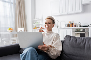 excited woman with trendy hairstyle laughing near laptop on sofa at home