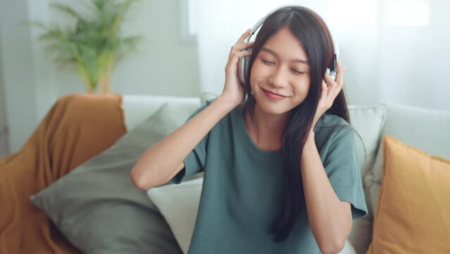 Happy young asian woman wear wireless headphones having fun listen music in smartphone and sit on sofa in living room, Female enjoys listening chill music audio sound feeling no stress at home. slow m