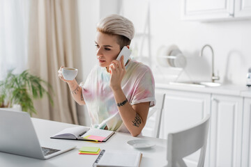 tattooed woman holding coffee cup during conversation on smartphone near laptop and notebooks