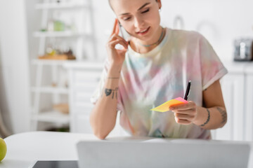 Obraz na płótnie Canvas positive woman holding sticky notes while talking on smartphone near laptop