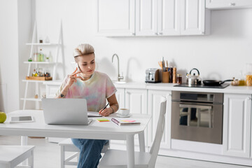 young woman talking on smartphone and writing in notebook near laptop in kitchen