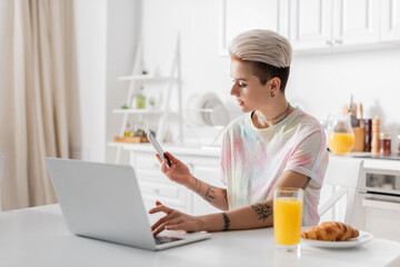 trendy tattooed woman with mobile phone typing on laptop near orange juice and croissant