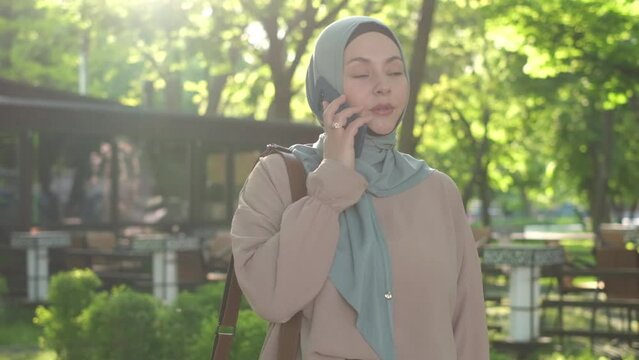 A Muslim woman walks in a park wearing a hijab and using a mobile phone. Smiling happy islamic girl talking on mobile phone