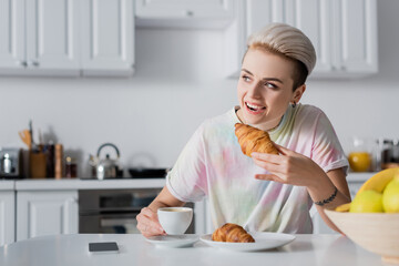 cheerful woman holding coffee cup and eating tasty croissant near smartphone with blank screen