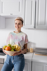 cheerful and stylish woman with bowl of ripe fruits smiling at camera in kitchen