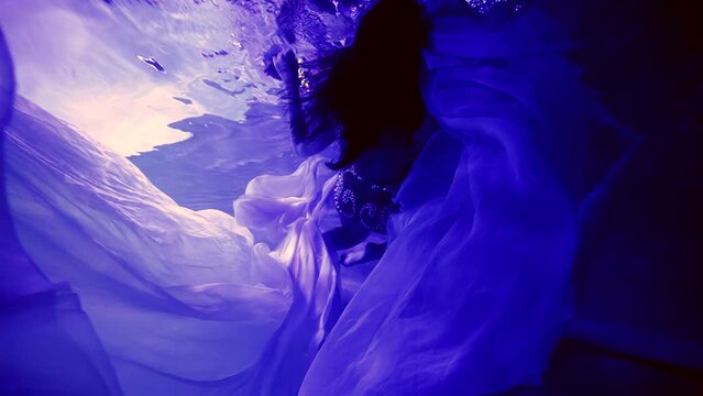 In the dark under the water, an elegant woman in a long dress swims like a mermaid in a fairy tale