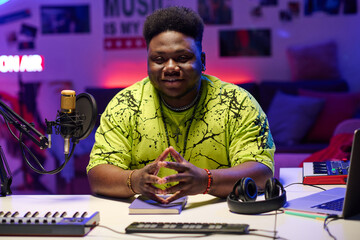 Stylish young Black man wearing neon yellow T-shirt sitting in at desk in amateur broadcasting...