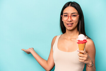 Young hispanic woman holding an ice cream isolated on blue background showing a copy space on a palm and holding another hand on waist.