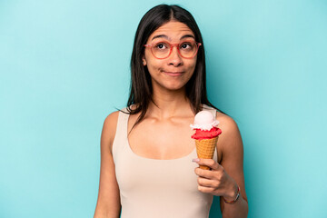 Young hispanic woman holding an ice cream isolated on blue background confused, feels doubtful and...