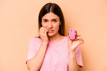 Young hispanic woman holding a keys car isolated on beige background biting fingernails, nervous and very anxious.