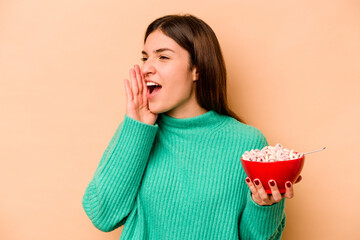 Young hispanic woman eating cereals isolated on beige background shouting and holding palm near opened mouth.