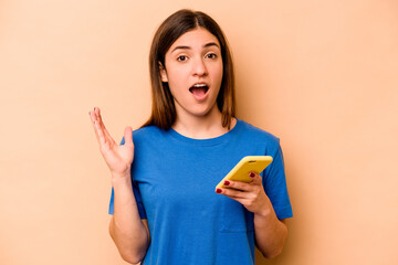 Young caucasian woman holding mobile phone isolated on beige background surprised and shocked.