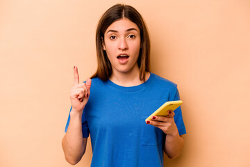 Young caucasian woman holding mobile phone isolated on beige background having an idea, inspiration concept.