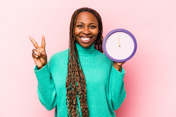 Young African American woman holding a clock isolated on pink background showing number two with...