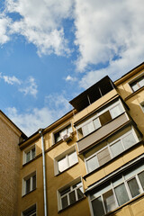 Low angle view of residential yellow house with balconies, windows and drainpipe. Blue sky with clouds. Air conditioner hangs on facade of house.