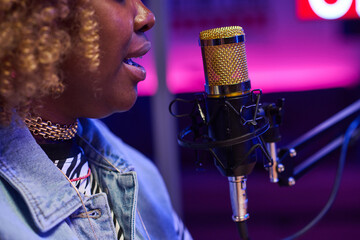 Side view close-up of young Black woman sitting in front of microphone recording podcast or doing...
