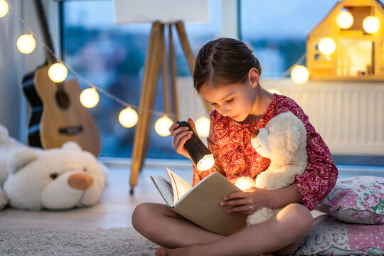 Beautiful brown hair toddler girl reads a book with flashlight in the bedroom. Child sitting with Teddy bear and fairy tale before bedtime. Christmas lights in children bedroom