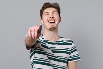 Fototapeta premium Young laughing fun cool caucasian man 20s wear blue striped t-shirt point index finger camera on you mocking joking kidding isolated on plain gray background studio portrait. People lifestyle concept.