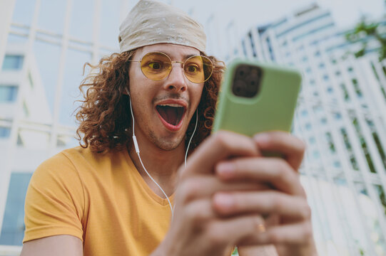 Young surprised amazed man 20s in yellow t-shirt bandana talk speak on mobile cell phone listen to music in headphones sitting rest relax in city outdoors on open air. Urban lifestyle leisure concept.