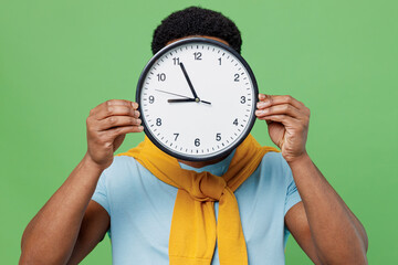 Young student brunet man of African American ethnicity 20s wearing blue t-shirt hold in hand clock cover face hiding isolated on plain green color background studio portrait. People lifestyle concept.